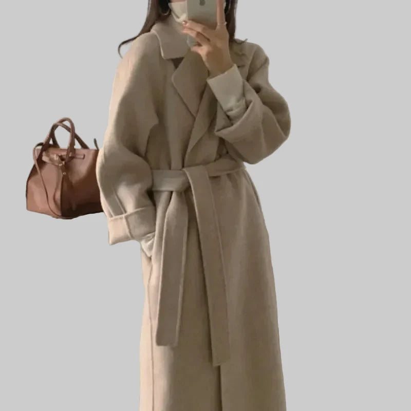 Kobieta w eleganckim beżowym długim płaszczu z paskiem w talii trzyma torbę