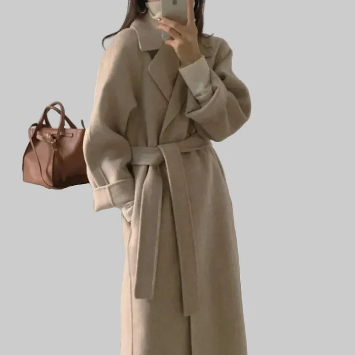 Kobieta w eleganckim beżowym długim płaszczu z paskiem w talii trzyma torbę